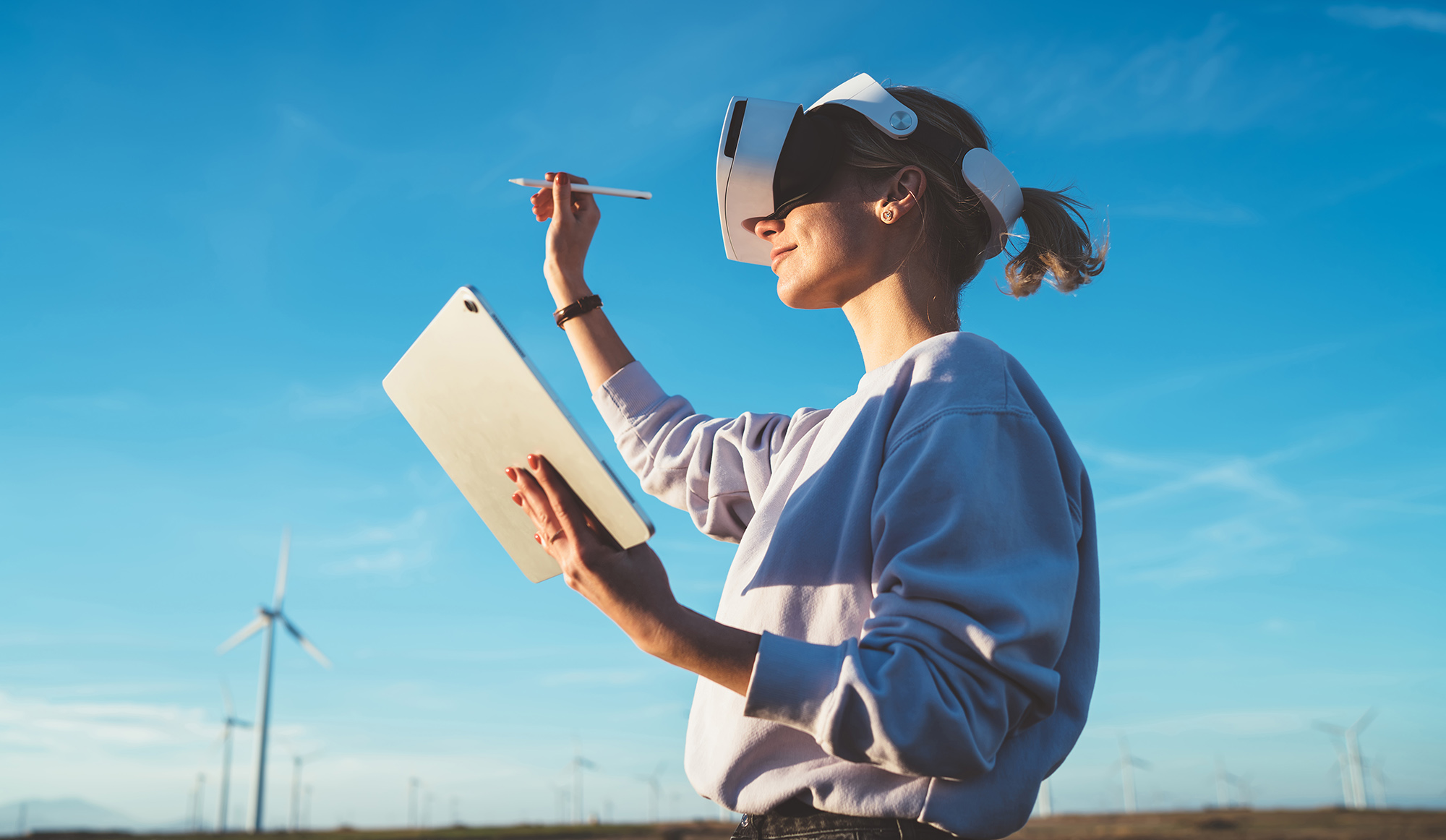 Blonde female in VR goggles making digital virtual reality space with touchpad program and stylus opposite sunlight on blue sky background with windmills